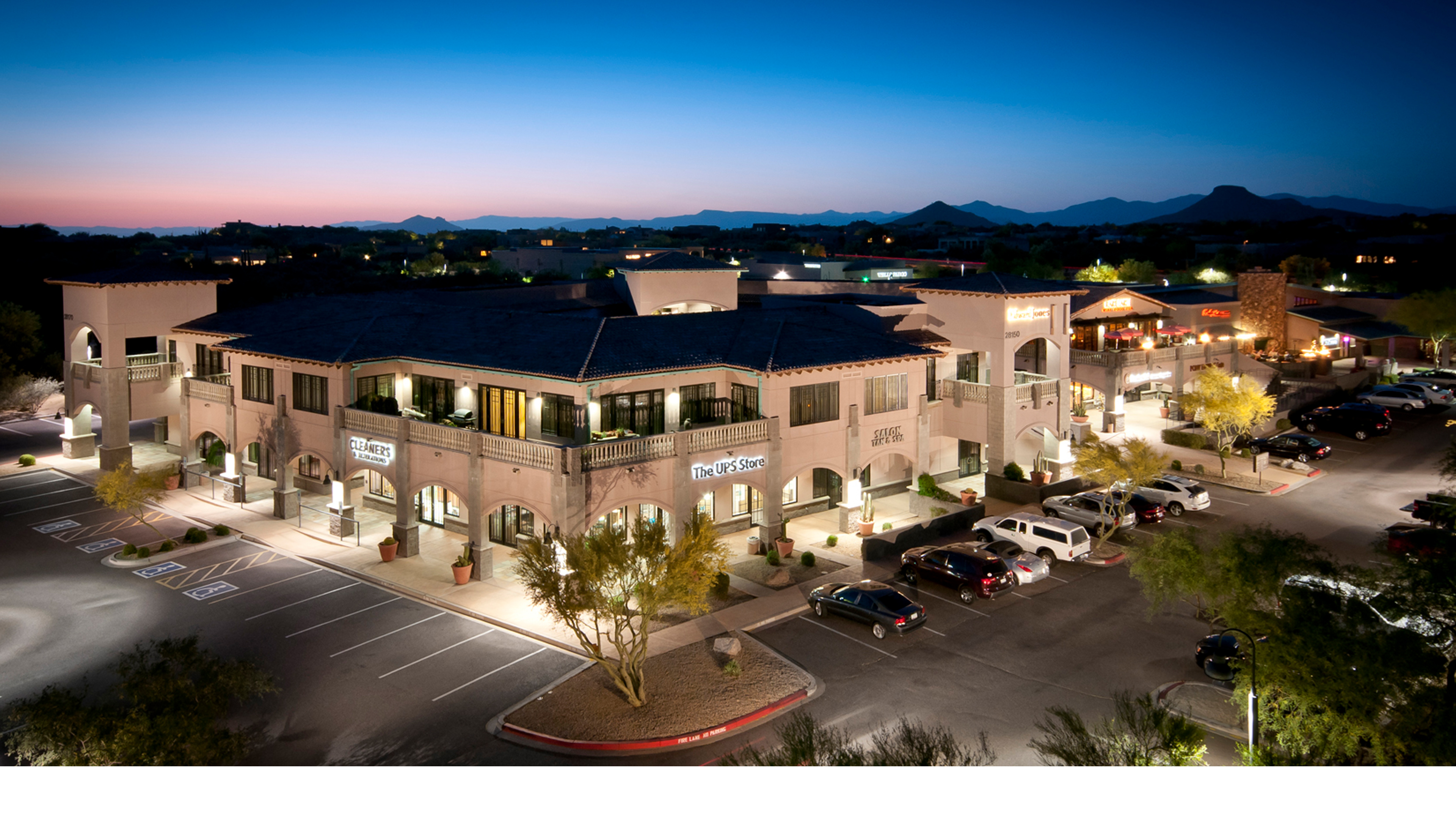 Center at Troon North retail and commercial development in Scottsdale, Arizona, illuminated at dusk with storefronts, parking and desert surroundings.